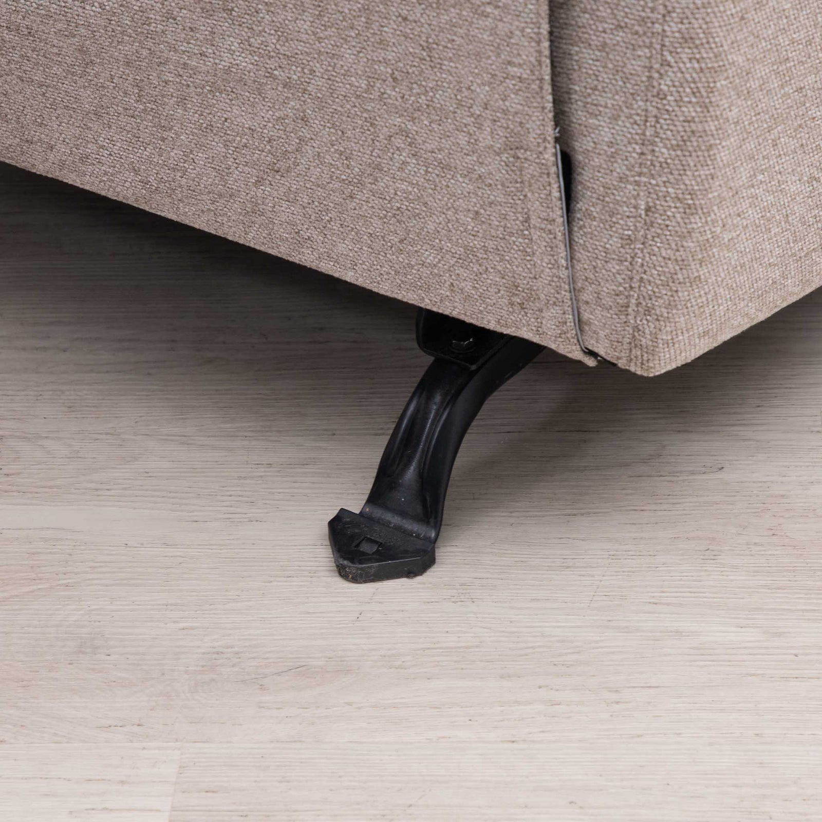 Close-up of the corner of a beige upholstered sofa with a black metal leg, resting on a light wood floor.