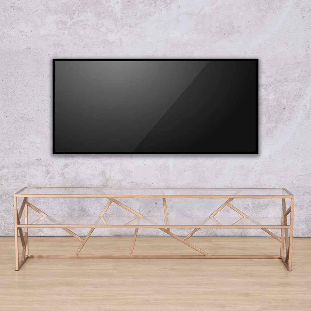 Front-facing view of the Legacy 1.8m TV Unit with a TV mounted, showcasing its gold mild steel base and clear glass top. | Leather Gallery