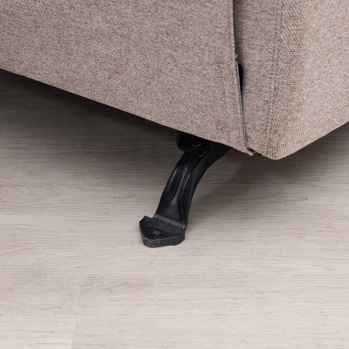 Close-up of the corner of a beige upholstered sofa with a black metal leg, resting on a light wood floor.