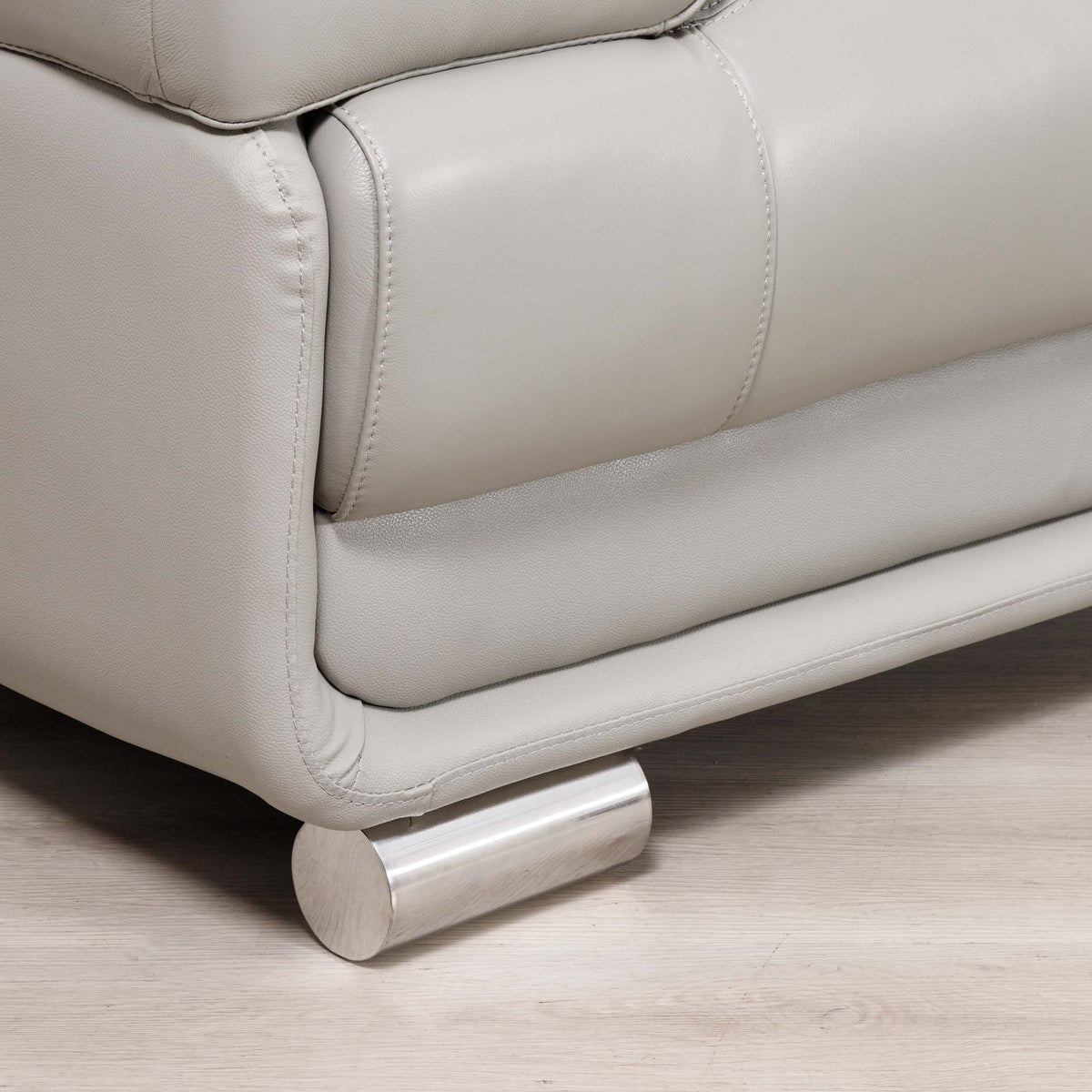 Close-up of a light gray leather sofa with a cylindrical brushed metal footrest, set on a light wood floor.