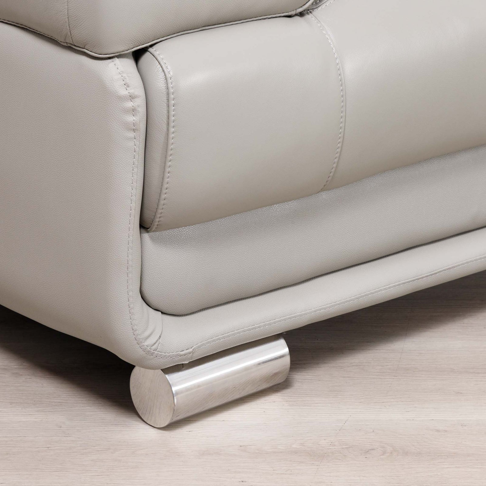 Close-up of a light gray leather sofa with a cylindrical brushed metal footrest, set on a light wood floor.