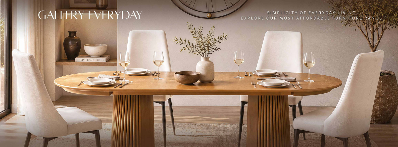 A modern dining room scene featuring a light oak wood oval dining table with fluted pedestal legs, surrounded by four white upholstered dining chairs. The table is set with white plates, wine glasses, and a central vase with dried branches.