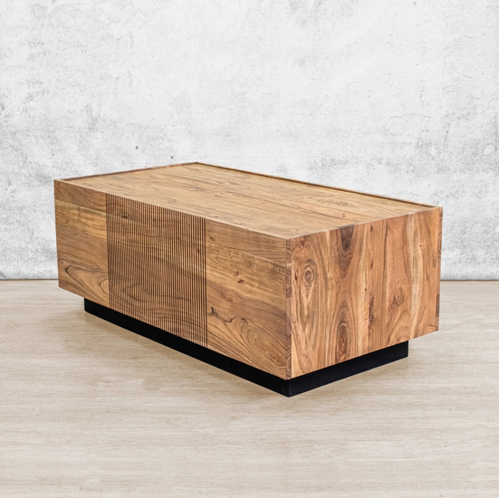 Angled view of a rectangular wood coffee table featuring a black plinth base and a decorative slatted center panel. 