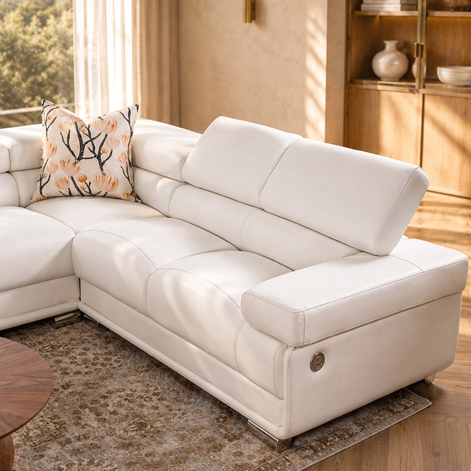 A modern white leather sectional sofa with a chaise lounge on the left, styled in a warm, sunlit living room with a patterned accent pillow and a textured rug.